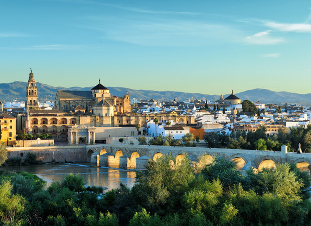 A historic stone bridge spans a river in front of a grand, ancient cathedral with a cityscape and mountains in the background under a clear blue sky.